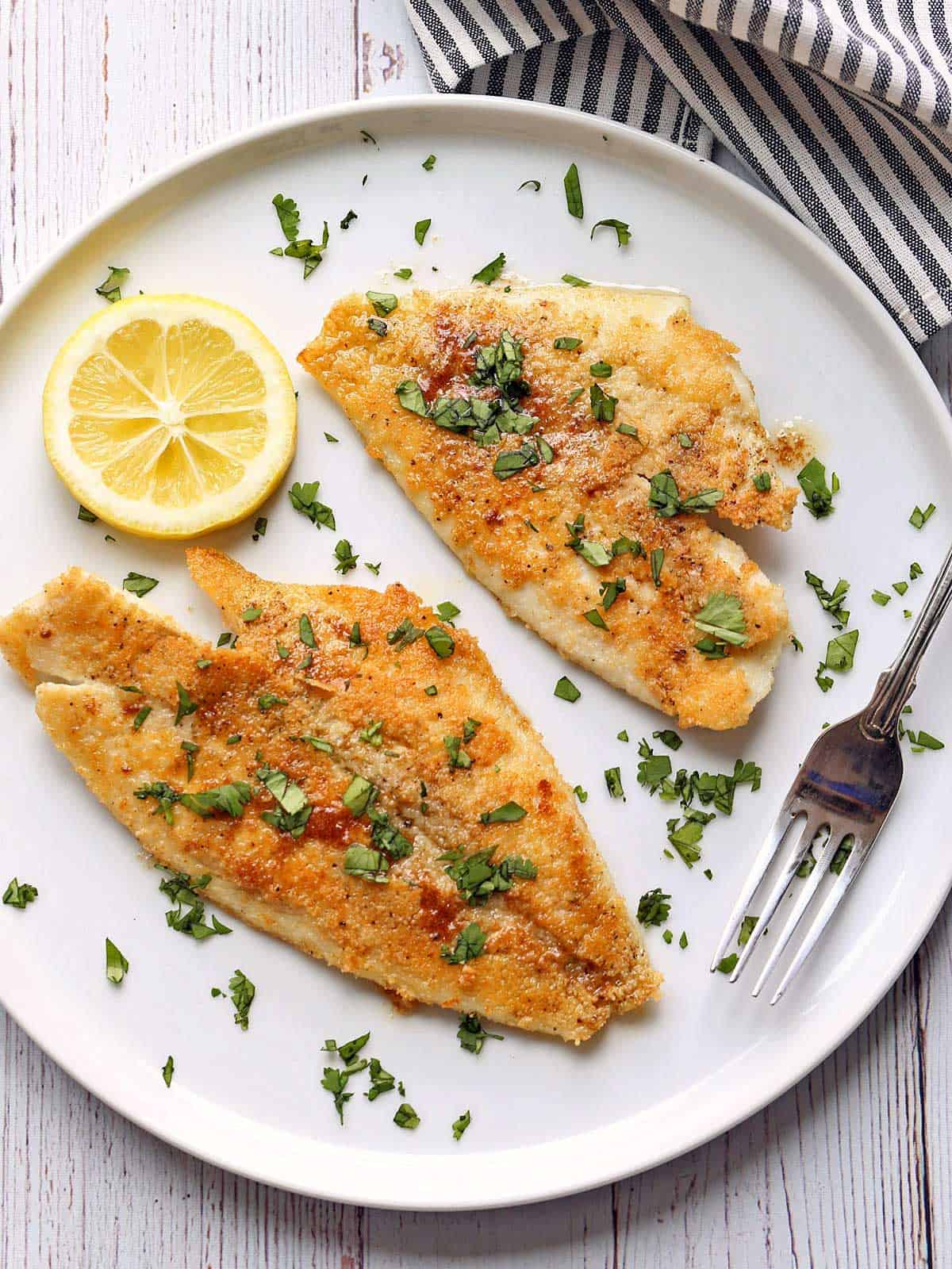 Two pieces of sole meuniere are served on a white plate with  a slice of lemon.
