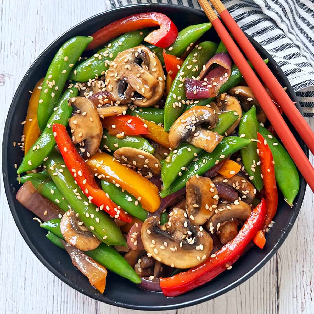 A vegetable stir-fry is served in a black bowl with red chopsticks.